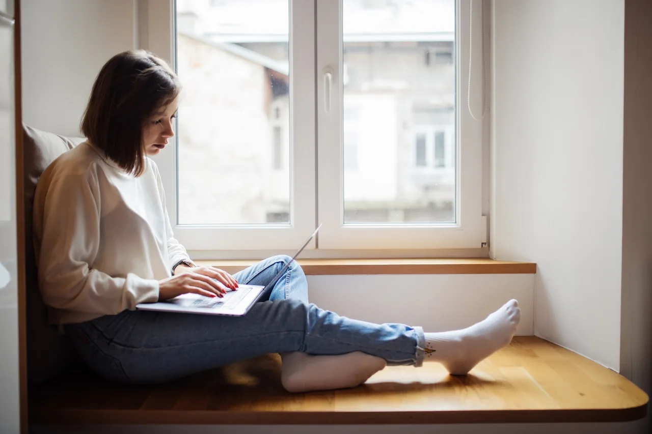 Žena s notebookem sedí na širokém dřevěném parapetu u okna